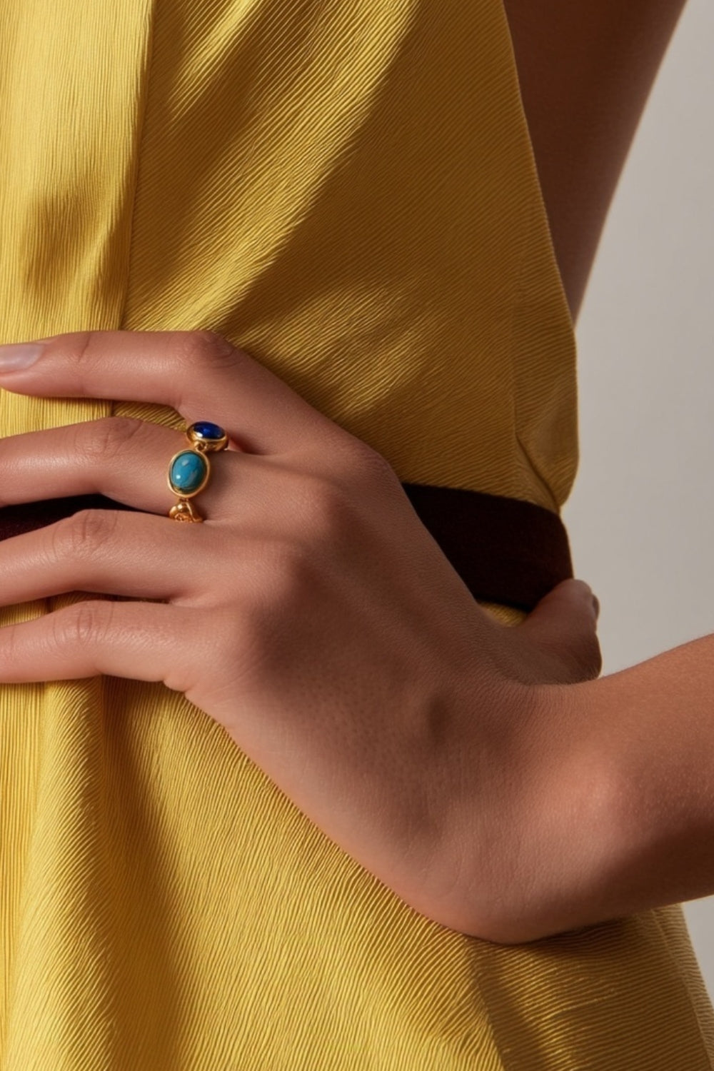 Close-up of a model's hand wearing a multicolor gemstone yellow gold ring with a yellow garment in the background