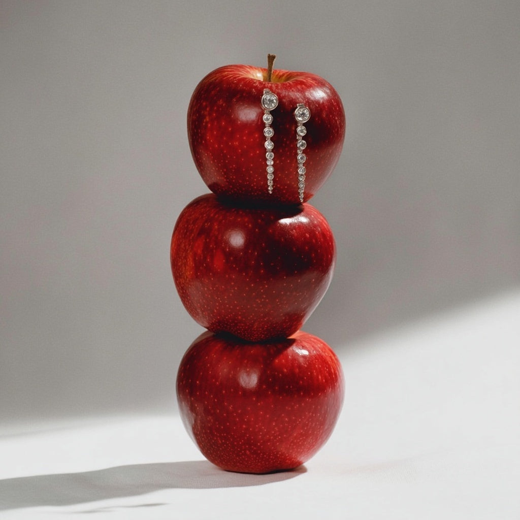 Three red apples stacked on top of each other with a pair of earrings between them on a light gray background.